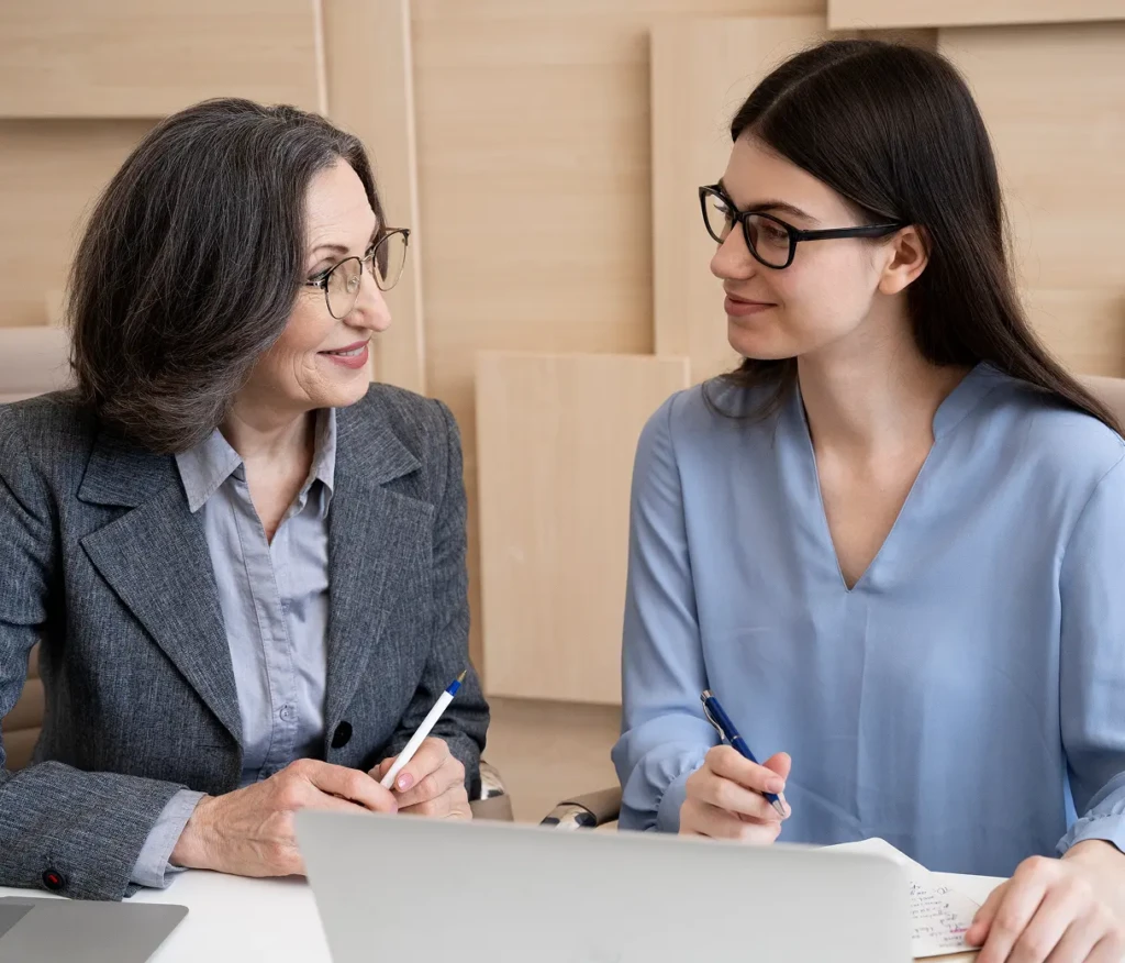 Two women sit at a table, smiling and holding pens. One wears a grey suit, the other a blue blouse. They appear engaged and collaborative.