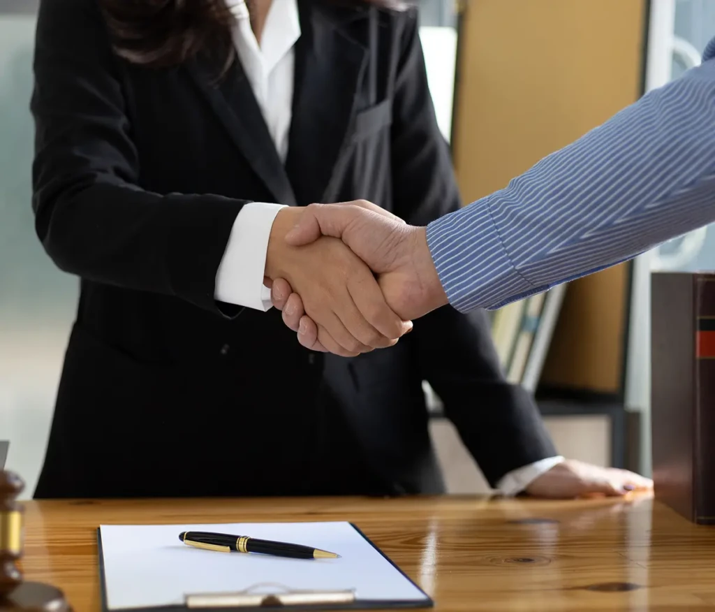 Two people shaking hands over a wooden desk with a contract and pen. One wears a black suit, the other, a blue striped shirt. The tone is professional.