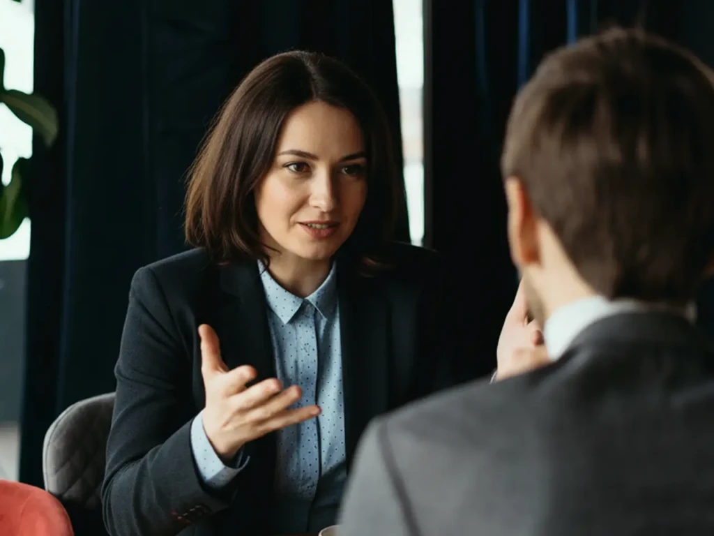 A woman in a suit gestures while speaking to a man in a business setting. The atmosphere is professional and engaged, suggesting a serious discussion.